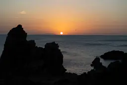 The setting sun above the sea and silhouetted coastal rocks.